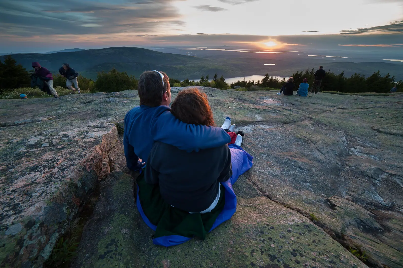 While seated on a blanket on a patch of mountaintop granite, a man and woman clasp their arms around